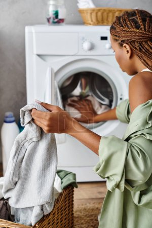 Eine afroamerikanische Frau mit Afro-Zöpfen legt in einem schön dekorierten Badezimmer fleißig Kleidung in einen modernen Trockner.