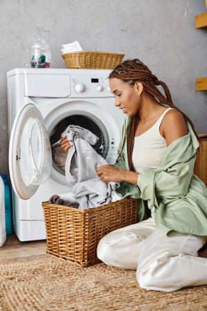 An African American woman with afro braids sitting on the floor next to a washing machine, doing laundry in a bathroom.