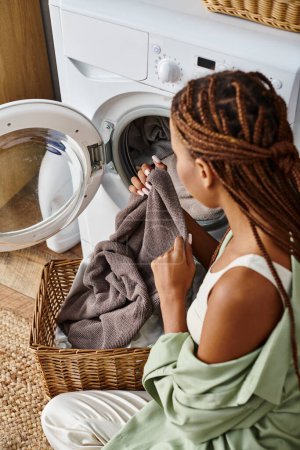 African American woman with afro braids sits next to a washing machine, doing laundry in a bathroom.