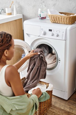 An African American woman with afro braids carefully loads clothes into a washing machine in a bathroom.