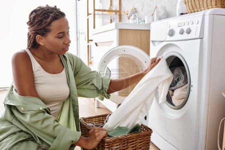 An African American woman with afro braids is doing laundry, putting clothes into a washing machine in a bathroom.