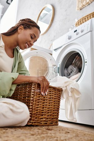 Téléchargez les photos : Femme afro-américaine avec des tresses afro assis sur le sol à côté de la machine à laver, faire la lessive dans la salle de bain. - en image libre de droit