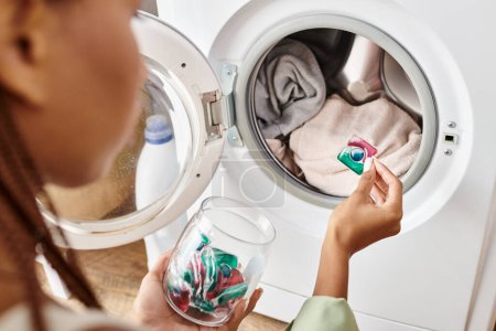 Une Afro-Américaine avec des tresses afro regarde la capsule de gel dans une machine à laver tout en faisant la lessive dans une salle de bain.