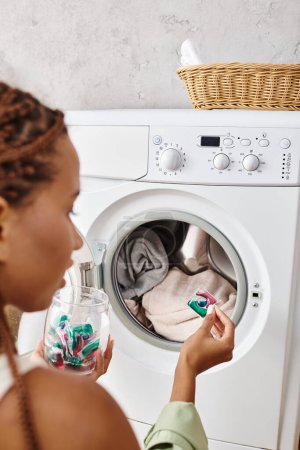Eine afroamerikanische Frau mit Afro-Zöpfen blickt in eine Waschmaschine im Badezimmer, während sie Wäsche wäscht.