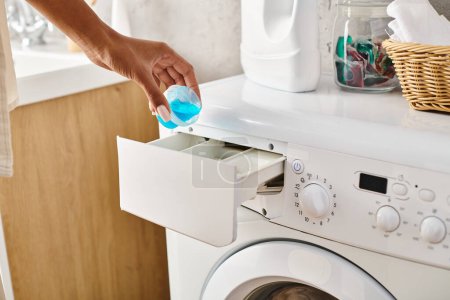 Una mujer afroamericana sostiene una cápsula de gel frente a una lavadora mientras lava la ropa en un baño.