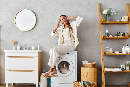 African American woman with afro braids comfortably sitting atop a washing machine while doing laundry in a bathroom.