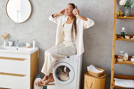 Mujer afroamericana con trenzas afro se sienta tranquilamente en una lavadora, lavando ropa en un baño.