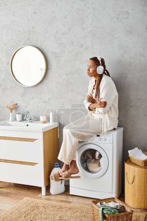 A woman with afro braids sits gracefully on a washing machine in a bathroom, doing laundry.