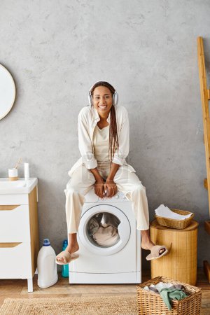 African American woman with afro braids sits on top of a washing machine while doing laundry in a bathroom.