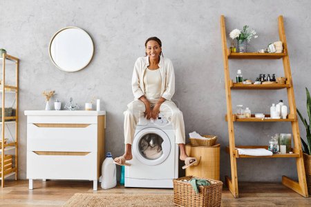 Photo for An African American woman with afro braids sitting on top of a washing machine, doing laundry in a bathroom. - Royalty Free Image