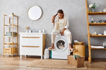 Téléchargez les photos : Femme afro-américaine avec des tresses afro se trouve au sommet d'une machine à laver, faire la lessive dans une salle de bain. - en image libre de droit