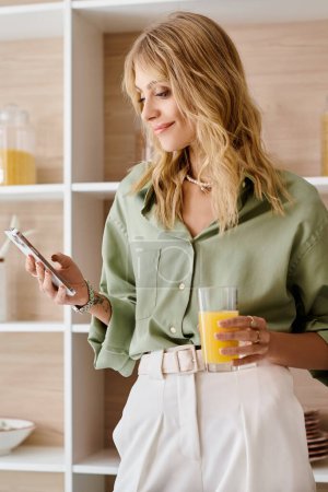 A woman standing in a kitchen holding a cell phone in one hand and a glass of orange juice in the other.