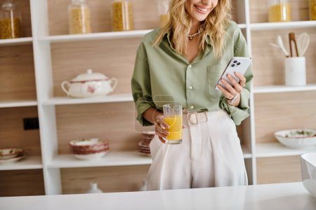 A woman standing in a kitchen holding a cell phone and a glass of orange juice.