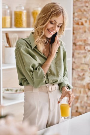 Una mujer en una cocina hablando en un teléfono celular mientras sostiene un vaso de jugo de naranja.