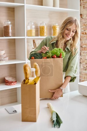 Woman standing in kitchen, holding a full bag of fresh vegetables.