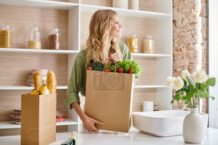 Téléchargez les photos : Une femme dans une cuisine tenant un sac d'épicerie rempli de légumes frais. - en image libre de droit