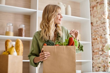 Une femme debout dans une cuisine, tenant un sac d'épicerie rempli de légumes.