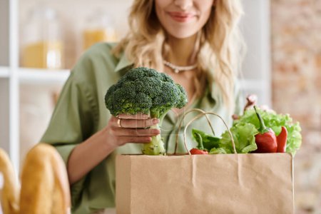 A woman holding a paper bag filled with various fresh vegetables in a kitchen.