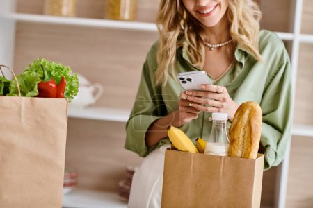 Eine Frau in der Küche blickt auf ihr Handy, während sie eine Tüte mit Lebensmitteln in der Hand hält.
