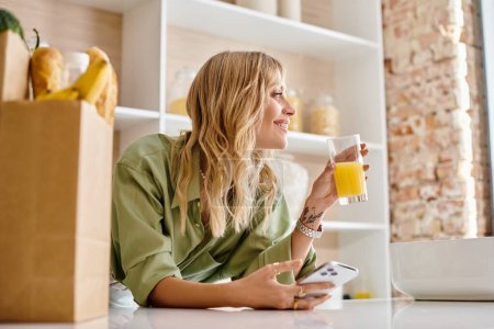 Téléchargez les photos : Une femme s'assoit à une table de cuisine tenant un verre de jus d'orange. - en image libre de droit