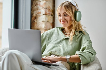 A woman sitting on a couch, wearing headphones, and using a laptop in a cozy apartment.