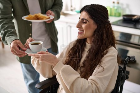 Téléchargez les photos : Femme handicapée gaie en fauteuil roulant tenant tasse de café à côté de son mari tandis que dans la cuisine - en image libre de droit