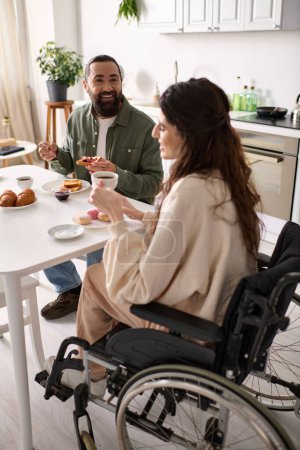 Photo for Joyful woman with inclusivity in wheelchair eating sweets at breakfast with her handsome husband - Royalty Free Image