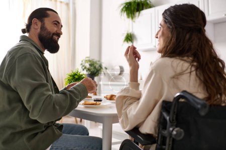 Téléchargez les photos : Beau joyeux homme appréciant le petit déjeuner avec sa femme heureuse handicapée en fauteuil roulant au petit déjeuner - en image libre de droit