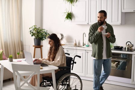 Téléchargez les photos : Bel homme buvant du café à côté de sa femme handicapée joyeuse en fauteuil roulant travaillant à l'ordinateur portable - en image libre de droit