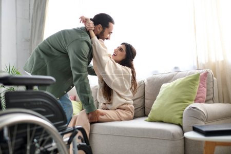 loving man in homewear helping his wife with mobility disability to get to couch from wheelchair