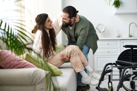 Liebender Mann in Hauskleidung hilft seiner gehbehinderten Frau, vom Rollstuhl auf die Couch zu gelangen