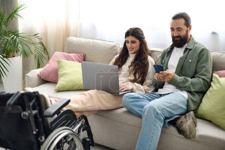 hermosa mujer con discapacidad de movilidad sentado en el sofá con el ordenador portátil junto a su marido con teléfono