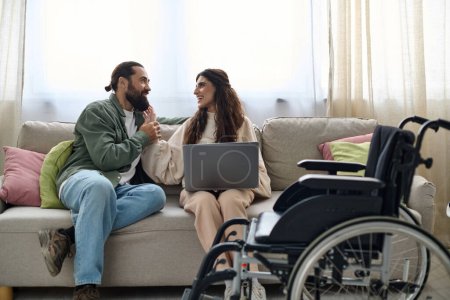 Foto de Alegre hombre mirando a la computadora portátil al lado de su discapacitada hermosa esposa alegre mientras está sentado en el sofá en casa - Imagen libre de derechos
