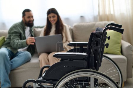 Foto de Se centran en la silla de ruedas moderna frente al hombre barbudo borroso y la mujer discapacitada mirando a la computadora portátil - Imagen libre de derechos