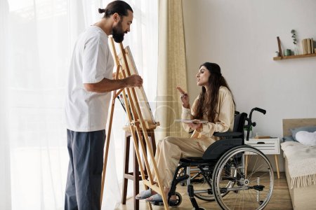 schöner bärtiger Mann beobachtet seine behinderte schöne Frau im Rollstuhl beim Bemalen auf der Staffelei zu Hause