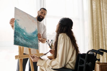 Photo for Beautiful woman with mobility disability painting on easel next to her cheerful bearded husband - Royalty Free Image