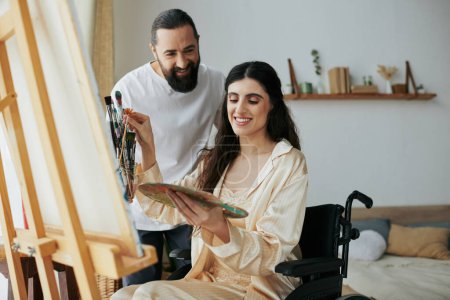 Foto de Atractivo barbudo hombre viendo su discapacitado hermosa esposa en silla de ruedas pintura en caballete en casa - Imagen libre de derechos