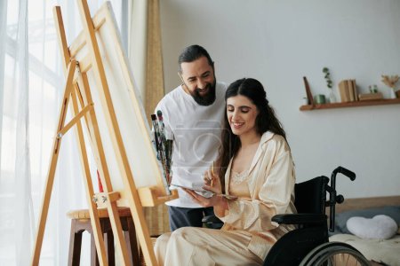 Foto de Atractivo barbudo hombre viendo su discapacitado hermosa esposa en silla de ruedas pintura en caballete en casa - Imagen libre de derechos