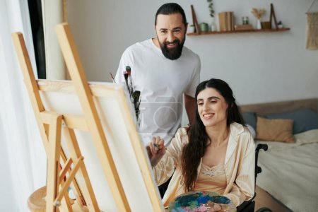 atractivo barbudo hombre viendo su discapacitado hermosa esposa en silla de ruedas pintura en caballete en casa