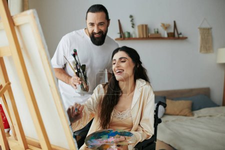 Téléchargez les photos : Aimant mari joyeux aidant sa belle femme inclusive à peindre sur chevalet pendant qu'il est à la maison - en image libre de droit