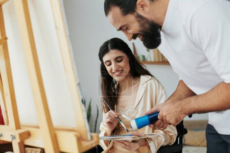 Photo for Loving jolly husband helping his inclusive good looking wife to paint on easel while at home - Royalty Free Image