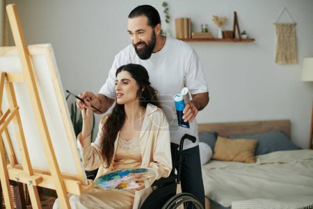 Téléchargez les photos : Soins joyeux mari aider son inclusive belle femme à peindre sur chevalet tout en à la maison - en image libre de droit