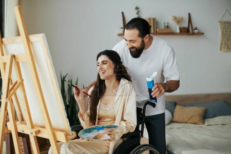 caring cheerful couple of bearded man and disabled woman painting on easel together at home