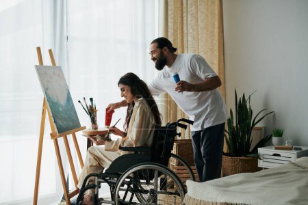caring cheerful couple of bearded man and disabled woman painting on easel together at home