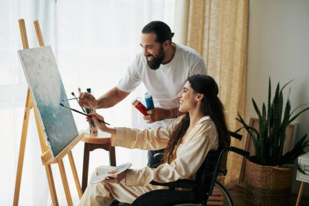 good looking bearded man watching disabled beautiful wife on wheelchair painting on easel at home