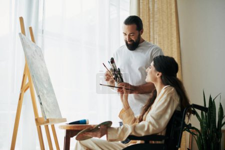 good looking bearded man watching disabled beautiful wife on wheelchair painting on easel at home