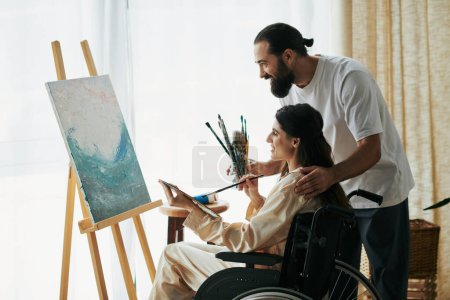 Foto de Atractiva pareja alegre de hombre barbudo y mujer discapacitada pintando en caballete juntos en casa - Imagen libre de derechos