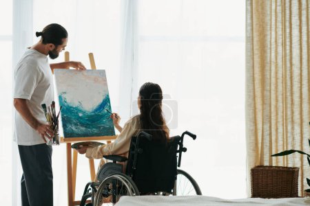 Téléchargez les photos : Attrayant couple joyeux de barbu homme et femme handicapée peinture sur chevalet ensemble à la maison - en image libre de droit