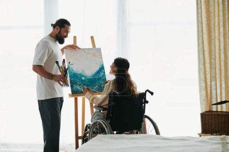 Photo for Attractive cheerful couple of bearded man and disabled woman painting on easel together at home - Royalty Free Image