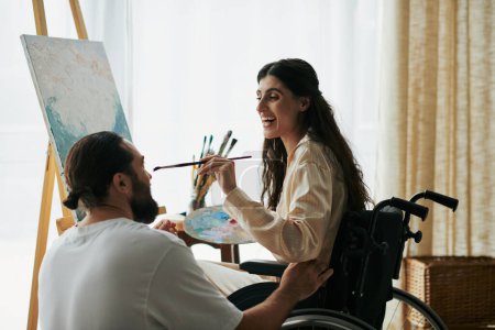 Foto de Marido positivo ayudando a su hermosa esposa inclusiva en silla de ruedas a pintar en caballete mientras está en casa - Imagen libre de derechos